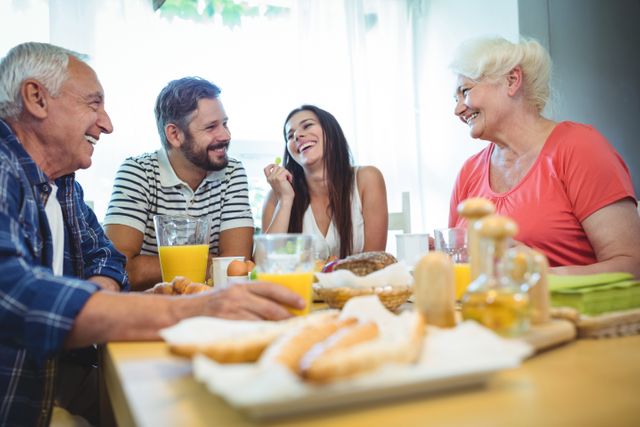 Older couple with their adult children enjoying breakfast together and laughing. Fresh food and beverages, including orange juice, on the table. Perfect for illustrating family time, intergenerational bonding, homely atmosphere, and joyful moments.