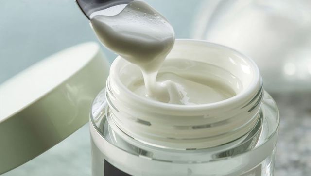 Scooping glossy white moisturizer cream with spatula above open glass jar macro studio