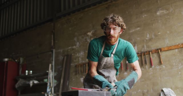 Caucasian male blacksmith wearing safety glasses, hammering hot metal tool on anvil in workshop. small business and craftsmanship at a blacksmiths forge.