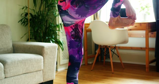 A person practices yoga at home in vividly colored leggings. Room has natural light, with furniture and house plants, creating a calm and peaceful environment. Ideal for use in websites, blogs, and ads focusing on home fitness, yoga practice, mindful living, and wellness.