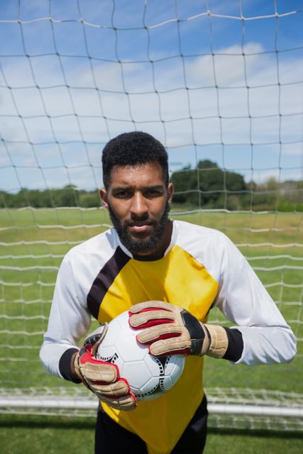 Goalkeeper standing in front of goal post holding football, wearing gloves and uniform. Ideal for use in sports-related content, advertisements for sports equipment, team promotions, and articles about soccer or athletic training.