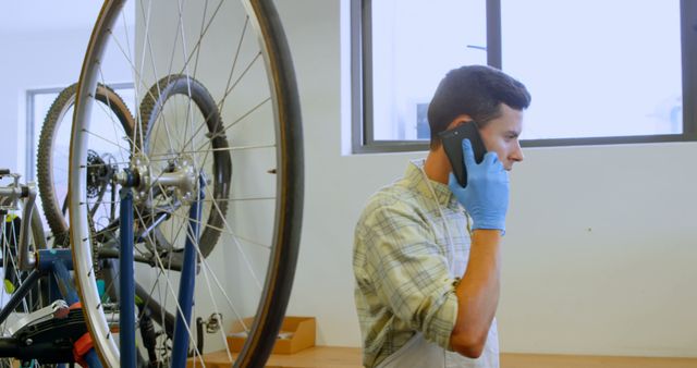 Bicycle mechanic standing in workshop answers phone call while wearing blue gloves. Bike rims visible in foreground, providing context of a professional repair setting. Useful for illustrating small businesses, repair shops, or customer service scenarios.