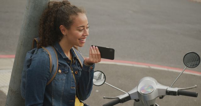 A young woman is talking on her smartphone while leaning against a pole next to an electric scooter. She is dressed in casual attire with a denim jacket and has a backpack. The scene depicts a modern urban lifestyle and can be used in themes related to communication, technology, transportation, and everyday activities.