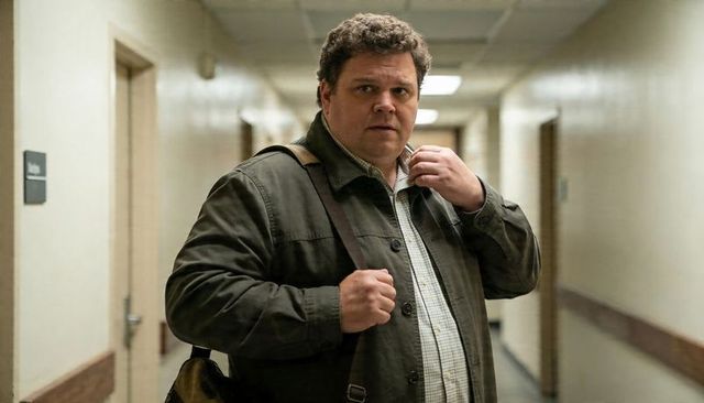 Middle-aged man adjusting jacket strap in narrow institutional hallway carrying leather satchel