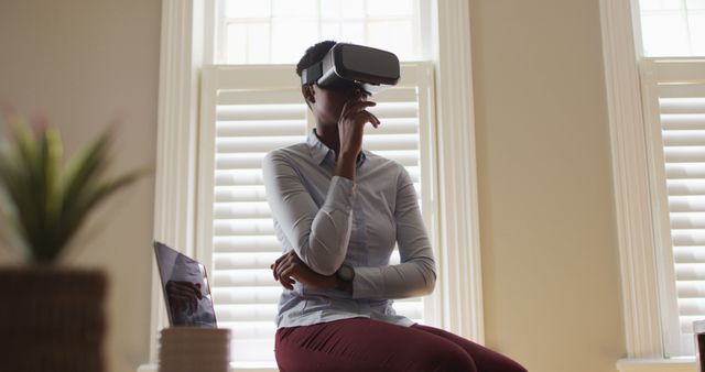 Professional woman wearing virtual reality headset sitting in modern daylight office. Surrounding includes large windows with blinds, creating a bright and well-lit ambiance. Perfect for illustrating the use of VR technology in professional settings, innovation in business, or modern office work environments.