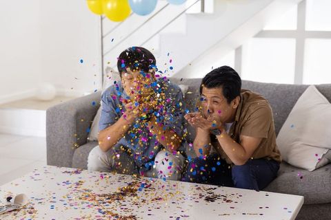 Asian men blowing colorful confetti on couch during playful indoor celebration with balloons