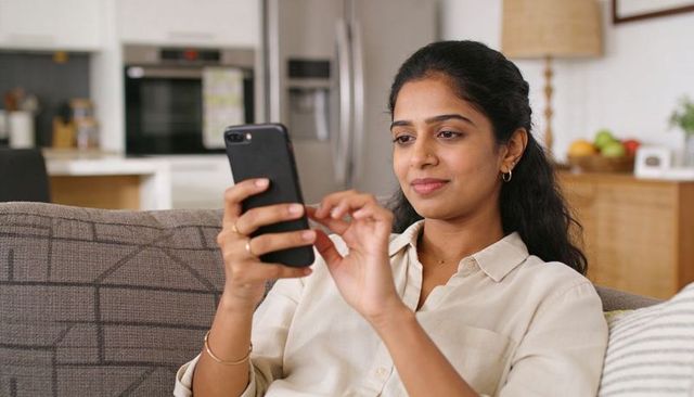 Indian woman scrolling smartphone on couch in modern open-plan kitchen relaxing at home