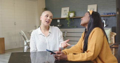 Diverse female friends chatting at home over smartphone, conversation in modern kitchen