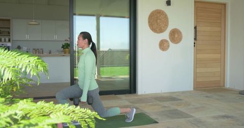 Asian woman lunging with dumbbells on patio on green mat doing home strength training