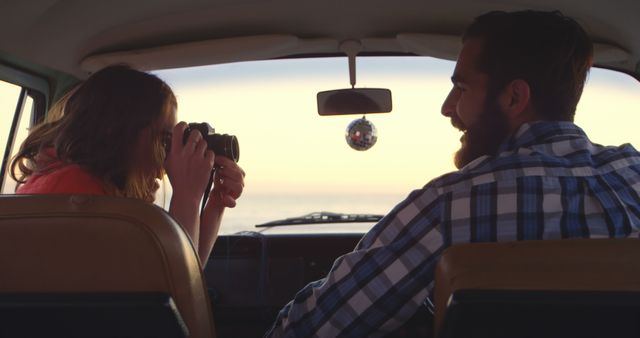 Couple is traveling in a camper van during sunset. The woman is taking photographs, while the man is smiling. Perfect for themes related to travel, adventure, vacations, love, and road trips.