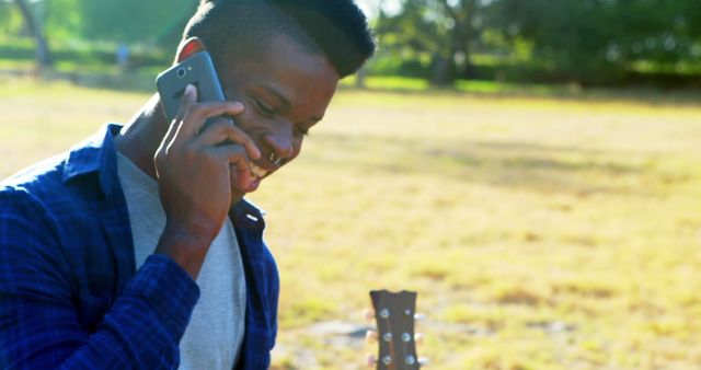 Young man smiling while talking on phone and holding a guitar in a park on a sunny day. Suitable for topics related to outdoor leisure, youth lifestyle, casual communication, music hobbies, and summer activities.