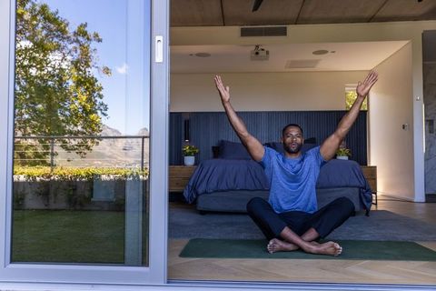 Mature man stretching arms while sitting cross-legged on yoga mat by open bedroom door