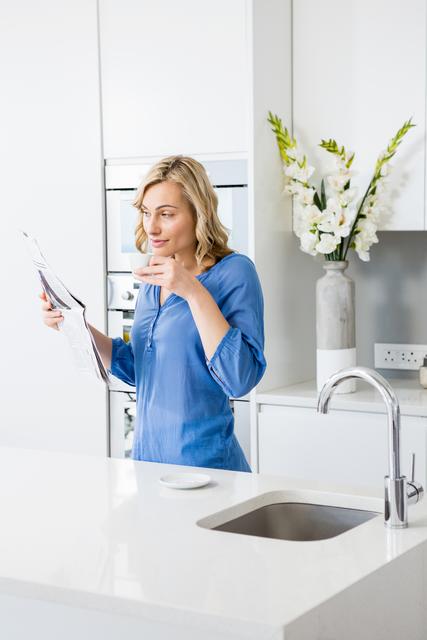 Woman in a modern kitchen holding a coffee cup and reading a newspaper. Ideal for use in lifestyle blogs, home decor magazines, and advertisements promoting morning routines, kitchen designs, or coffee brands.