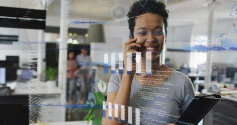 Businesswoman reviewing transparent data dashboards on glass while talking on phone