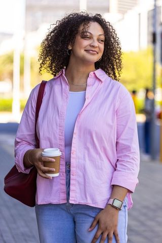 African american woman holding coffee cup, checking smartwatch and smiling on urban sidewalk