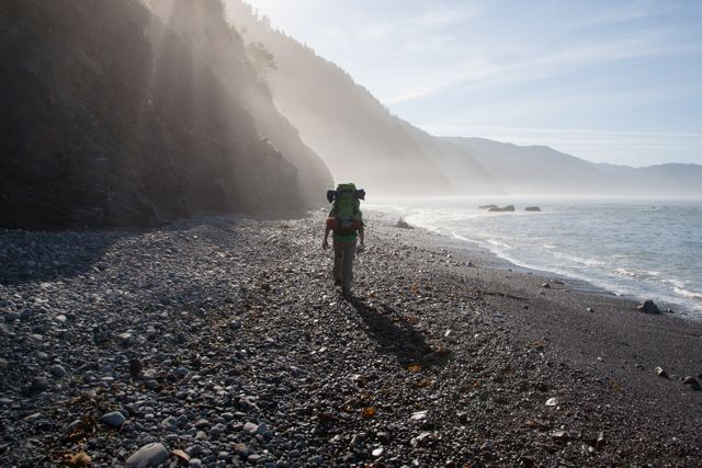 Backpacker walking along a rocky beach during sunrise with mist-covered hills in the background. Ideal for adventure-inspired articles, travel blogs, and nature lover websites.