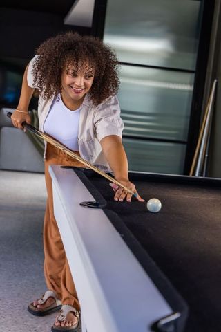 African american woman smiling and leaning over pool table aiming cue in modern game room