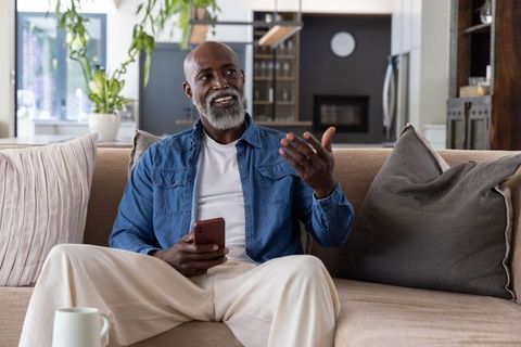 African american man sitting on sofa holding smartphone gesturing in modern living room