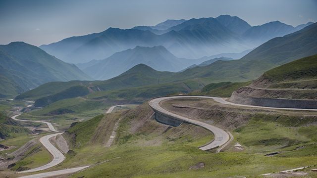 Captivating photo showing a winding road traversing through green hills on a misty day with distant mountains. Ideal for promoting outdoor adventures, travel destinations, scenic drives, and nature tourism. Also suitable for use in environmental documentaries or landscapes in publications.