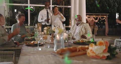 Couple toasting guests on porch at night around candlelit rustic wooden dinner table
