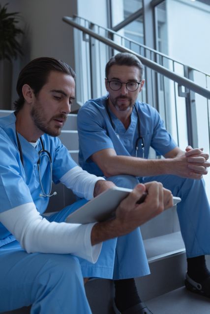 Two male surgeons in blue scrubs are sitting on stairs in a hospital, discussing patient records on a clipboard. This image can be used for healthcare and medical-related content, emphasizing teamwork, consultation, and professional collaboration in a hospital setting.
