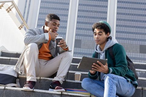 College students studying outdoors on urban steps using tablet and sipping coffee