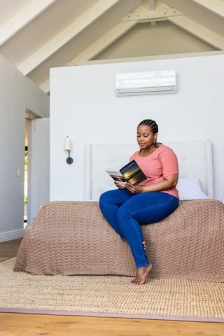 African american woman sitting on bed reading paperback in bright cozy minimalist bedroom