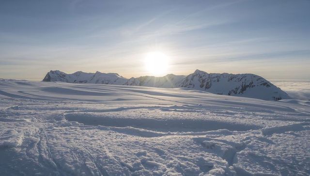 Sunlit Alpine Snowfield with Jagged Mountain Ridge at Sunrise on Wind-sculpted Plateau
