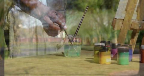 Mature Artist Mixing Paintbrushes in Outdoor Studio with Colorful Paint Jars and Easel