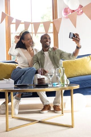 Couple taking selfie during home birthday celebration with cake, wine, balloons and tiara