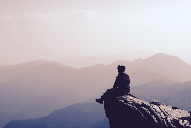 This image depicts a solitary man sitting on the edge of a cliff, overlooking an expansive, misty mountain range. The serene atmosphere and silhouette effect create a sense of peace and contemplation. This image is perfect for use in travel blogs, adventure-themed websites, nature and travel magazines, and in any media focusing on solitude, reflection, and the beauty of the natural world.