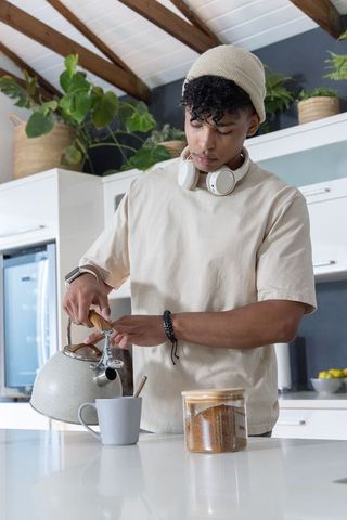 African american man pouring hot water into mug in modern home kitchen with headphones