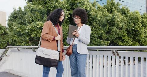 Female coworkers collaborating over tablet on urban bridge wearing blazers and jeans