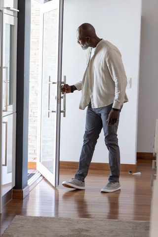 Senior african man opening glass front door in contemporary sunlit hardwood entryway