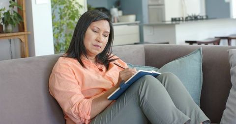 Mature asian woman writing in notebook on sofa at home, relaxed journaling lifestyle