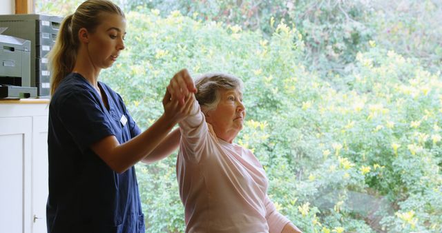 Young female caregiver is assisting an elderly woman with a mobility exercise in an outdoor garden setting. The caregiver is providing support by holding and guiding the woman's arm. The scene takes place in a peaceful and green garden, emphasizing healthcare and senior care. Perfect for use in healthcare, assisted living promotions, senior wellbeing articles, and caregiving support materials.