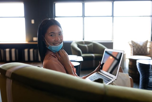 Asian woman wearing a face mask sitting on couch using laptop looking at camera smiling. health and hygiene in creative office during coronavirus covid 19 pandemic.