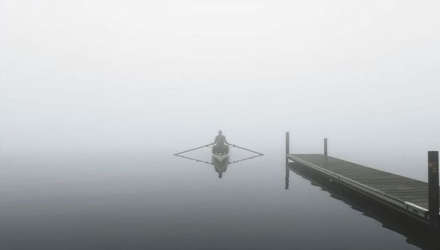 Lone rower in single scull gliding through dense fog beside wooden dock, minimalist reflection