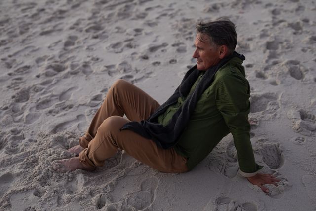 High angle side view of a happy senior Caucasian man enjoying time in nature, sitting on sand on a beach barefoot, admiring the view smiling relaxing.