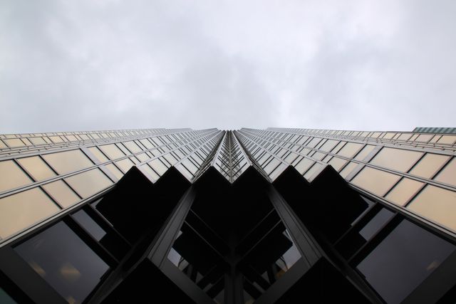 Abstract view of a modern skyscraper seen from below with a cloudy sky in the background. The glass facade and symmetrical lines create a geometric and futuristic perspective. Useful for articles on urban life, modern architecture, business districts, or corporate environments. Ideal for backgrounds, presentations, and web design emphasizing urban development and modern cityscapes.