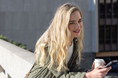 Blonde woman leaning on urban terrace railing smiling while holding coffee and smartphone