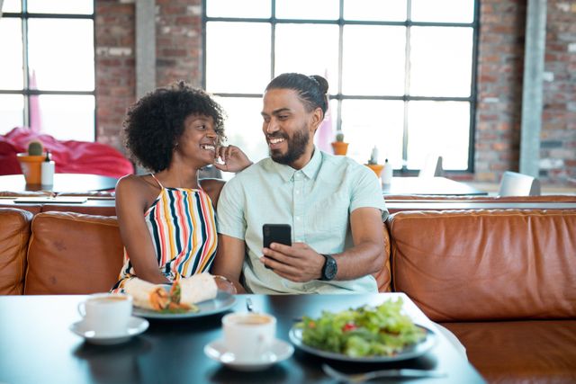 Smiling diverse couple having lunch in cafe looking at smartphone together. couple enjoying free time out together.
