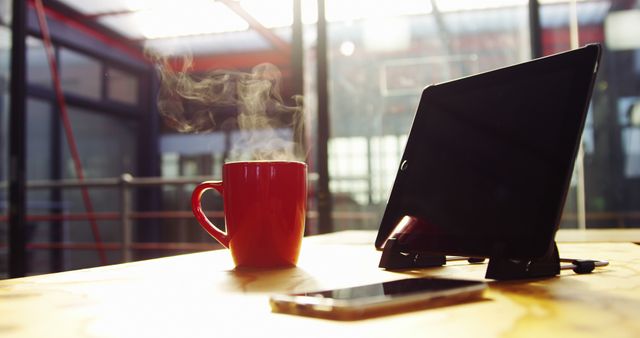 A steaming red coffee mug sits next to a smartphone and an open laptop on a wooden table, with copy space. Warm lighting and the cozy ambiance suggest a comfortable workspace for productivity or a casual meeting spot.
