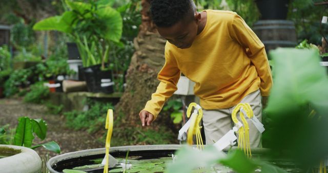 Young boy wearing yellow sweater, bending down to observe water features in lush outdoor garden, highlighting a moment of curiosity and connection with nature. Ideal for educational content about nature, promoting outdoor activities for children, or illustrating curiosity-driven designs.