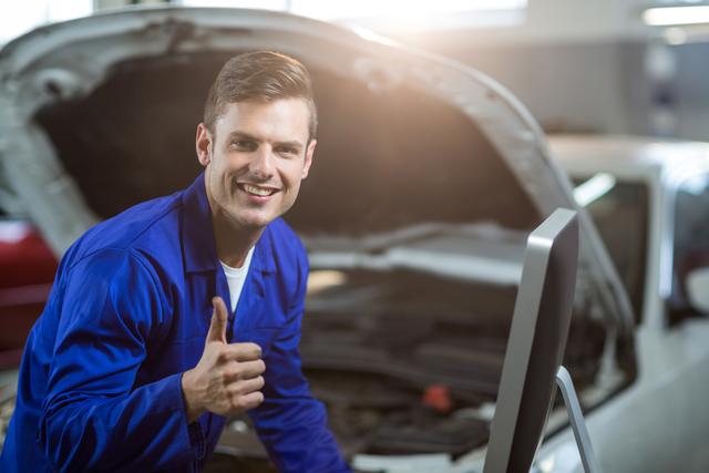 Mechanic in blue uniform smiling and showing thumbs up in front of car with open hood in garage workshop. Suitable for automotive service advertisements, repair shop promotions, and industry-related content.