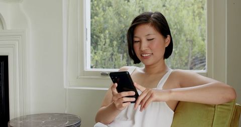 Asian Woman Relaxing at Home Using Smartphone Near Window