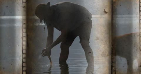 Backlit fisherman bending in shallow water holding small fish with vintage film border