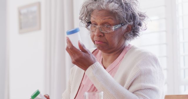 Senior African American woman with glasses holding and examining medicine bottles at home. Suitable for illustrating topics such as elderly care, medication management, health and wellness for aging populations, and independent living for seniors.