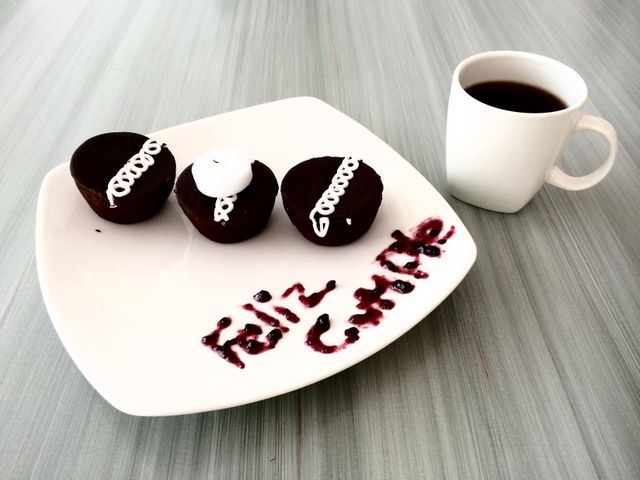 Three chocolate cupcakes with ornate white frosting sit on a white plate next to a cup of black coffee. The plate reads 'Feliz Cumple' in chocolate sauce, adding a festive touch. Ideal for birthday celebrations, dessert menus, marketing materials for bakeries, or social media posts about celebrations.