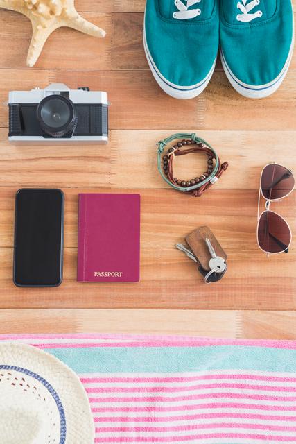 Various travel items and accessories arranged on a wooden surface. Includes a passport, a vintage camera, teal shoes, sunglasses, a smartphone, a bracelet, keys, and a striped towel. Perfect for blogs, social media posts, travel articles, and packing guides.